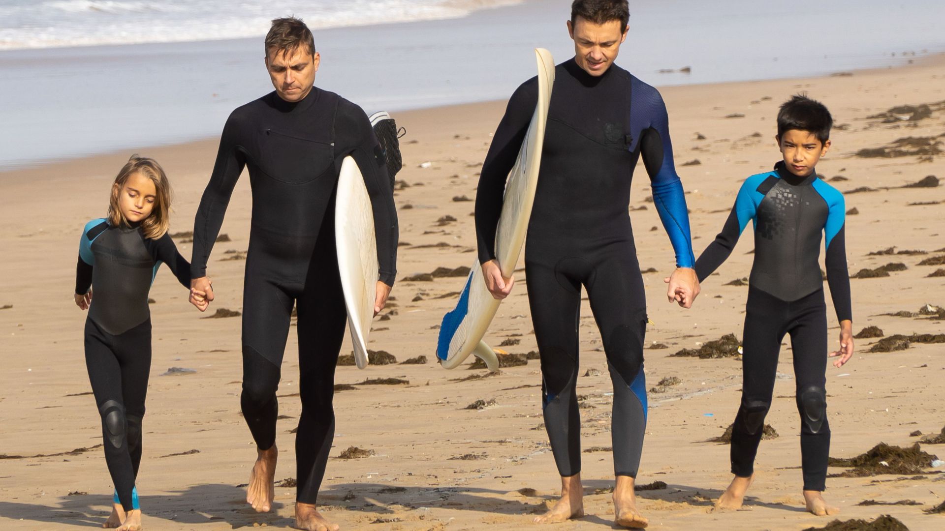 Two adults and two kids in wetsuits walking on a sandy beach in Portugal, carrying surfboards.