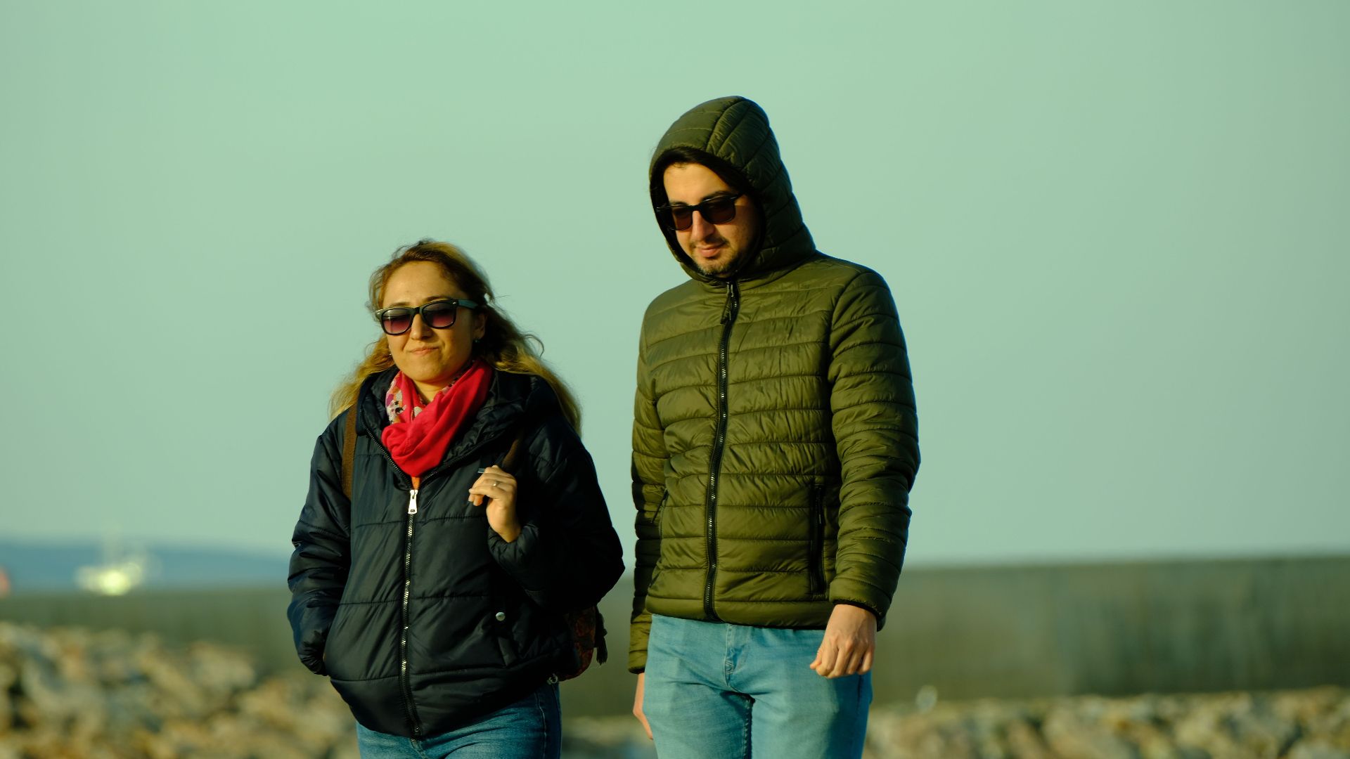A couple wearing winter jackets and sunglasses walking on a rocky beach.