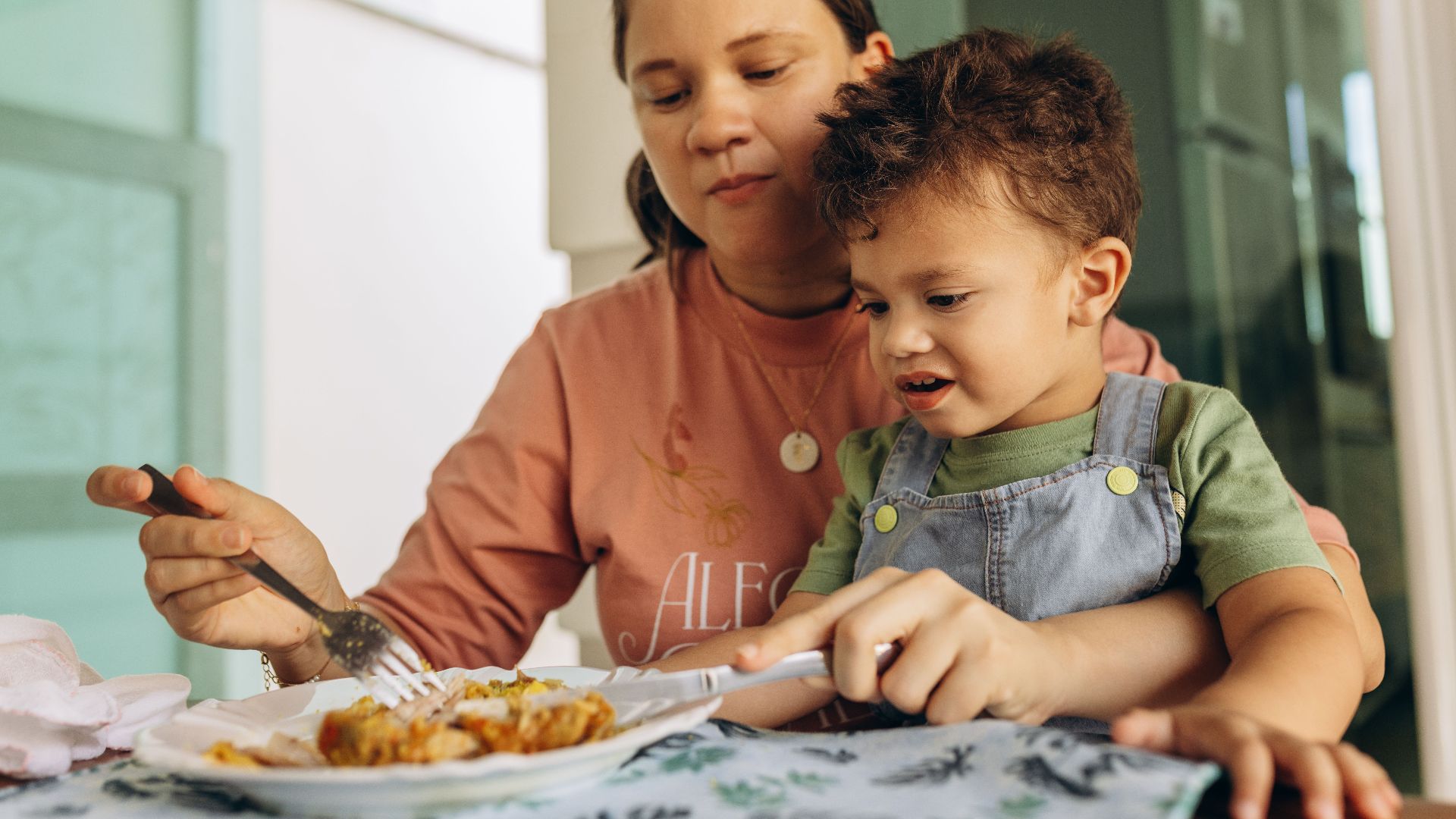 A mother helps her child eat at the kitchen table, fostering family bonding.