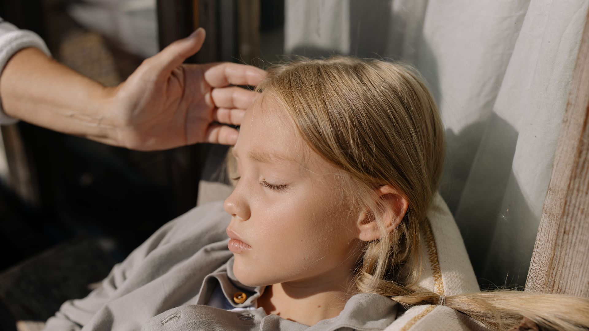 A resting child with eyes closed, gentle hand on forehead, captured in soft lighting.