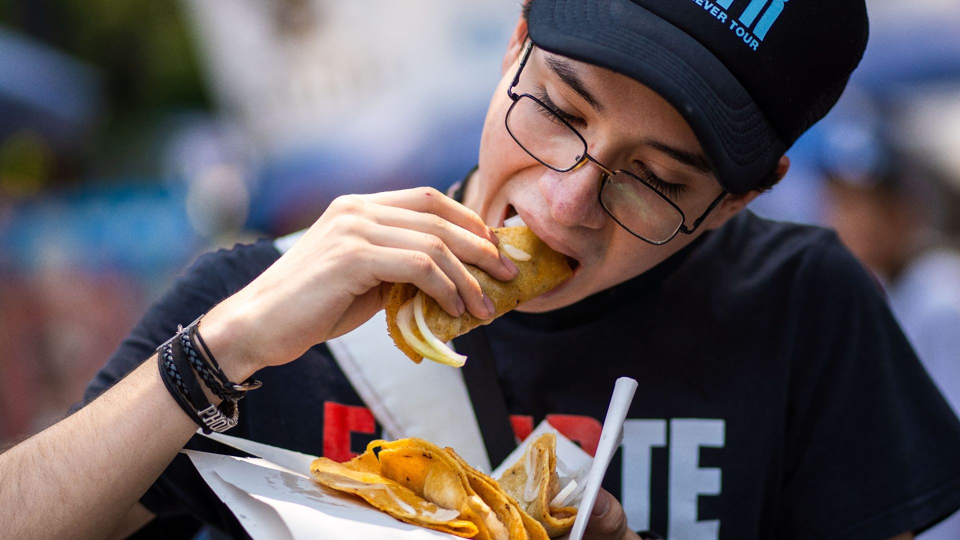 A young man wearing a cap and glasses enjoys street tacos in Mexico City.