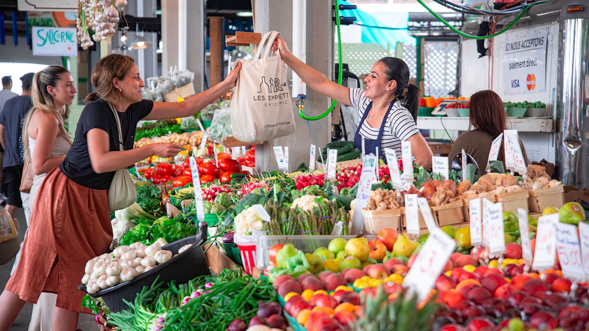 Women shopping at a colorful outdoor market stall filled with fresh fruits and vegetables. Smiles and interaction.