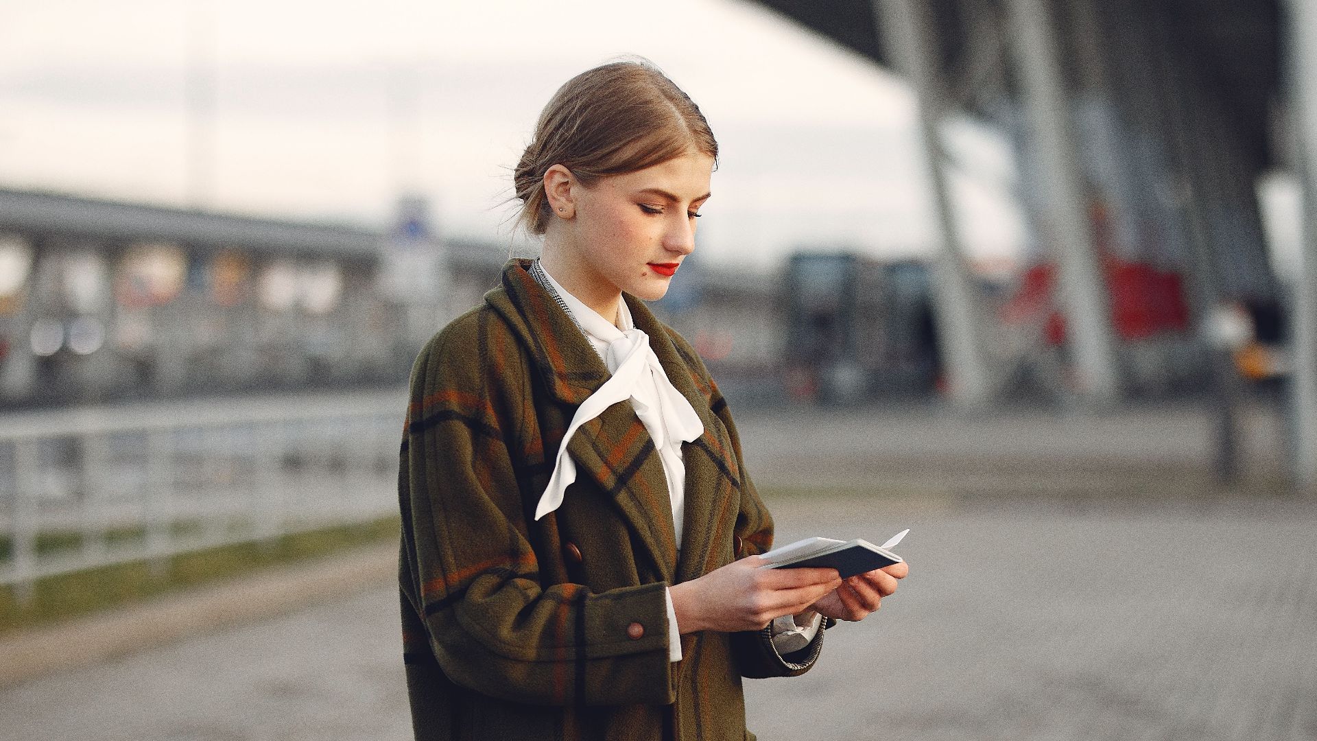 Attentive female passenger wearing trendy plaid coat and white blouse checking passport and ticket standing on pavement near modern building of airport outside
