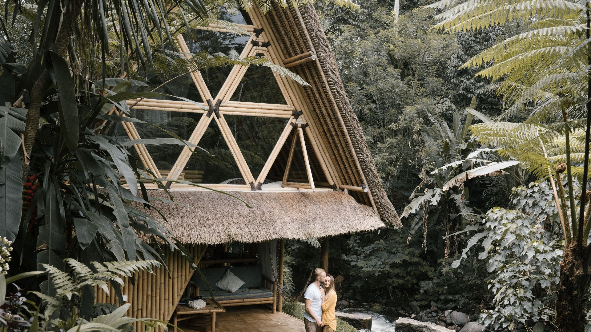 A couple embraces near a bamboo hut in a lush tropical garden by a gentle river.