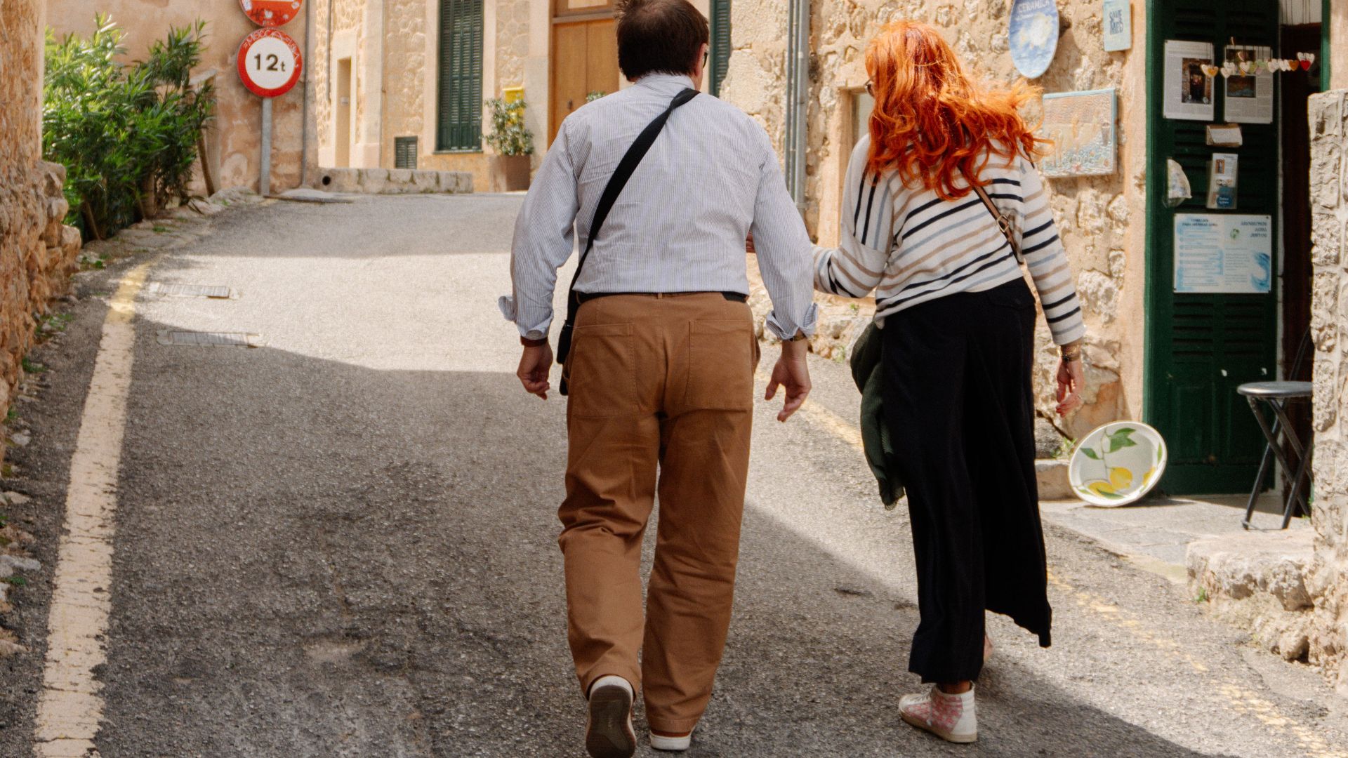 A couple walks through a picturesque Mediterranean village with stone buildings on a sunny day.