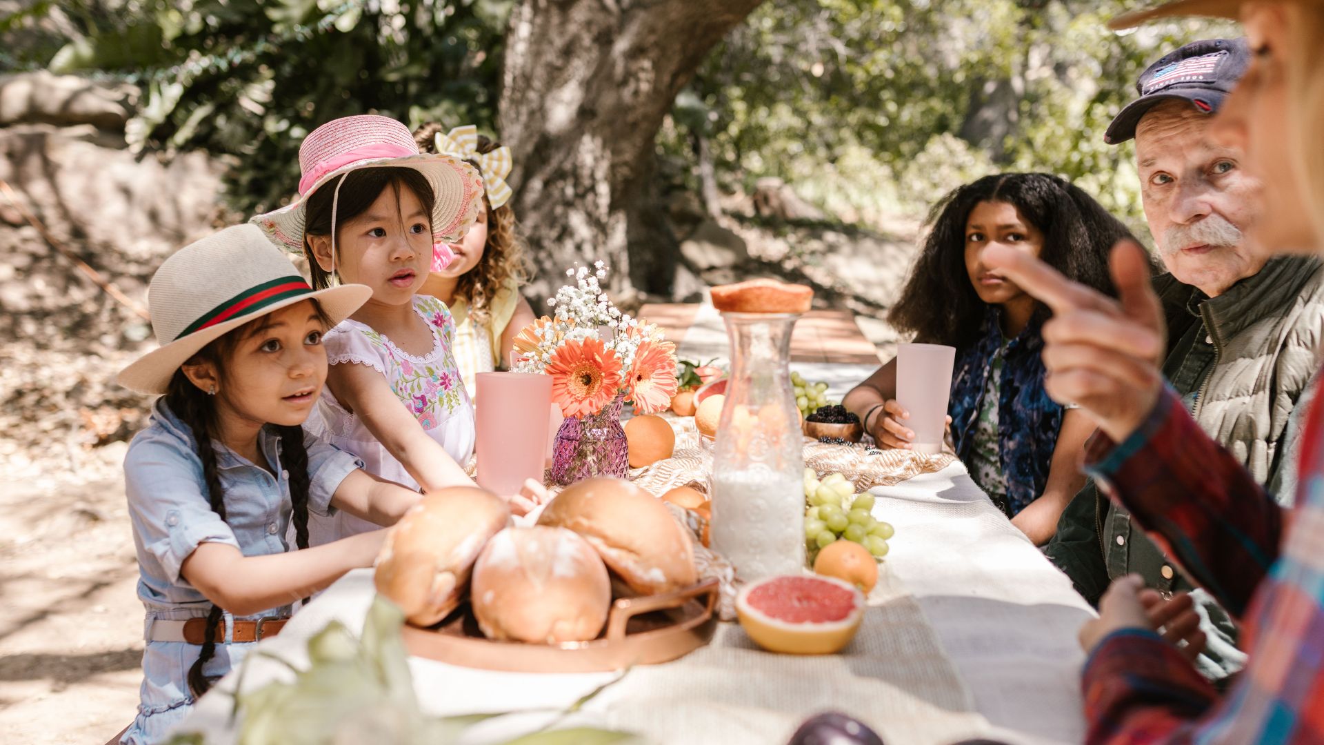 Diverse group gathered at an outdoor picnic table with breads and fruits on a sunny day.