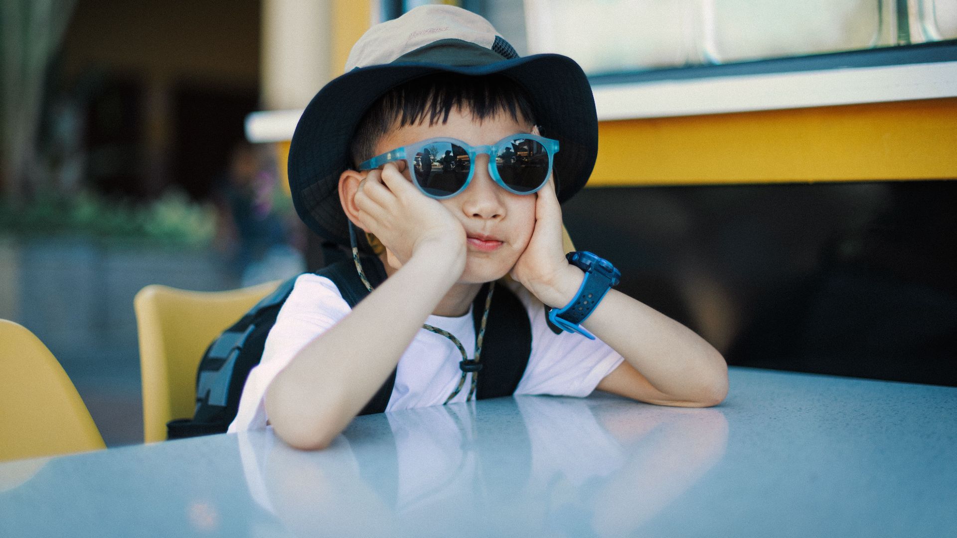 Child in sunglasses and hat sitting pensively at a table outdoors on a sunny day.