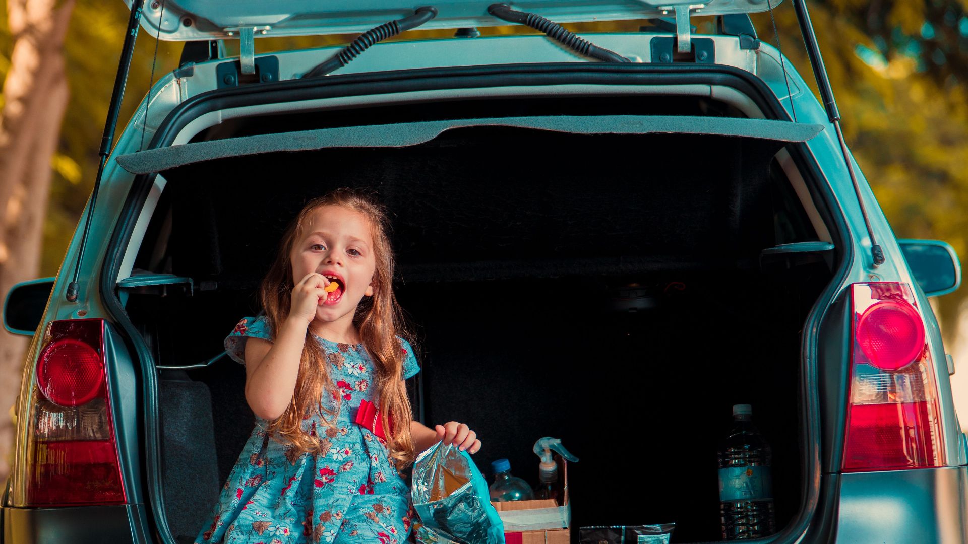 A young girl in a floral dress enjoys a snack sitting in the trunk of a car on a sunny day.