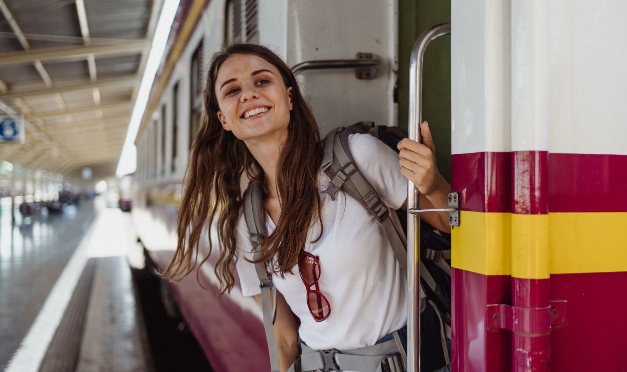 A cheerful woman with a backpack smiling while leaning out of a train at a station