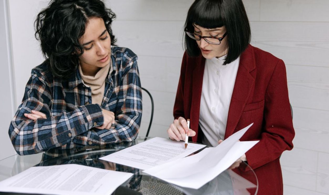 Two women collaborating at a glass table, reviewing business documents in a modern office setting