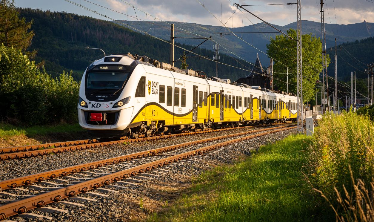 A modern yellow and white train travels through a picturesque countryside setting under a blue sky