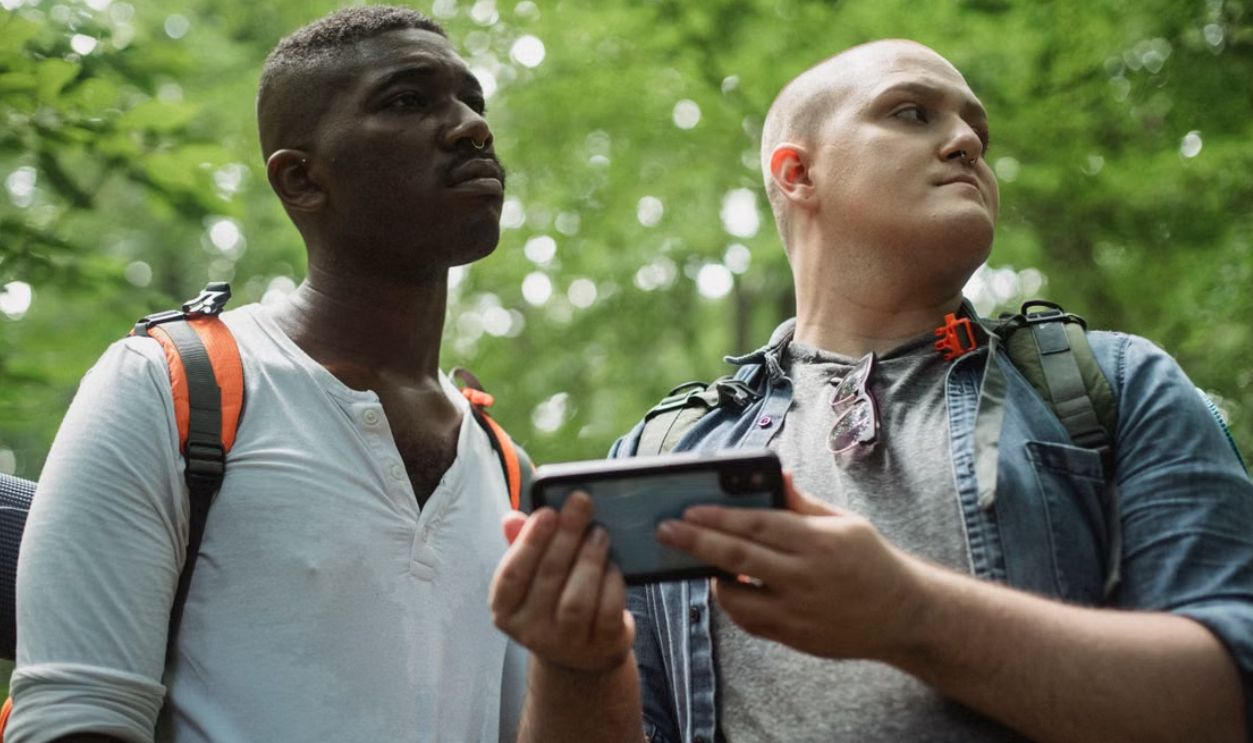 Two men using smartphones while hiking in a lush green forest, focused on navigation