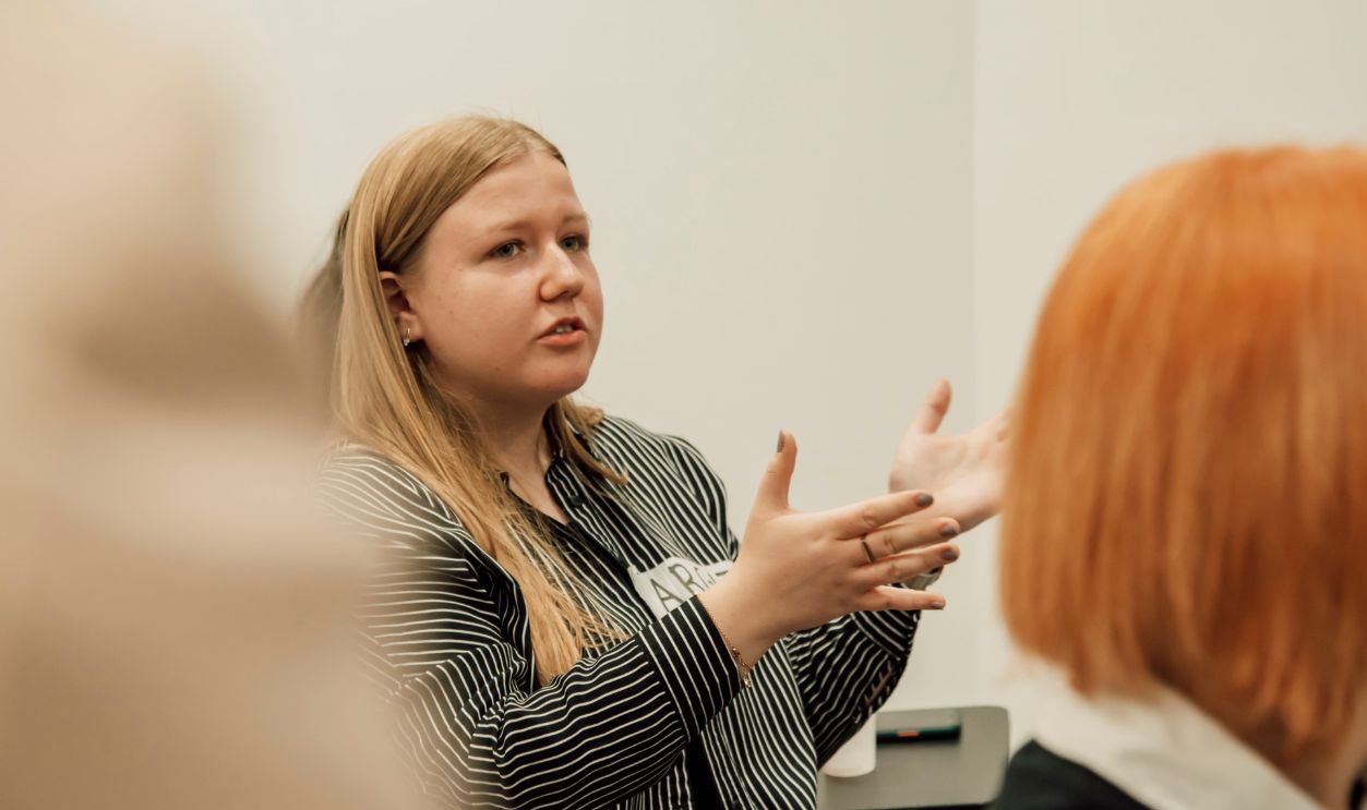 Woman with long blonde hair speaking passionately during a group meeting indoors