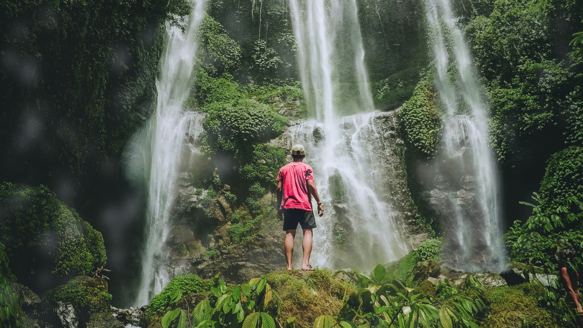 man in red shirt standing on rock near waterfalls during daytime