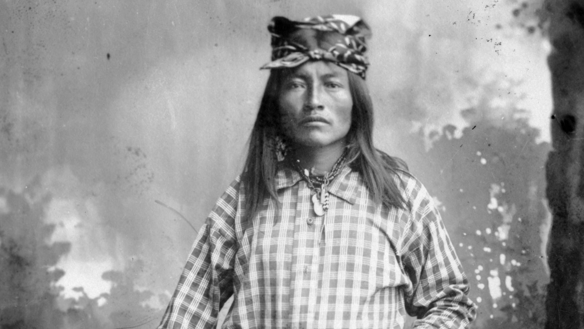 Studio portrait (standing) of Dutchy (Bakeitzogie [ Yellow Coyote]), a Native American (Chiricahua Apache) man. He holds a rifle and wears moccasin boots, a breechcloth, ammunition belt, and a kerchief on his head. Title and