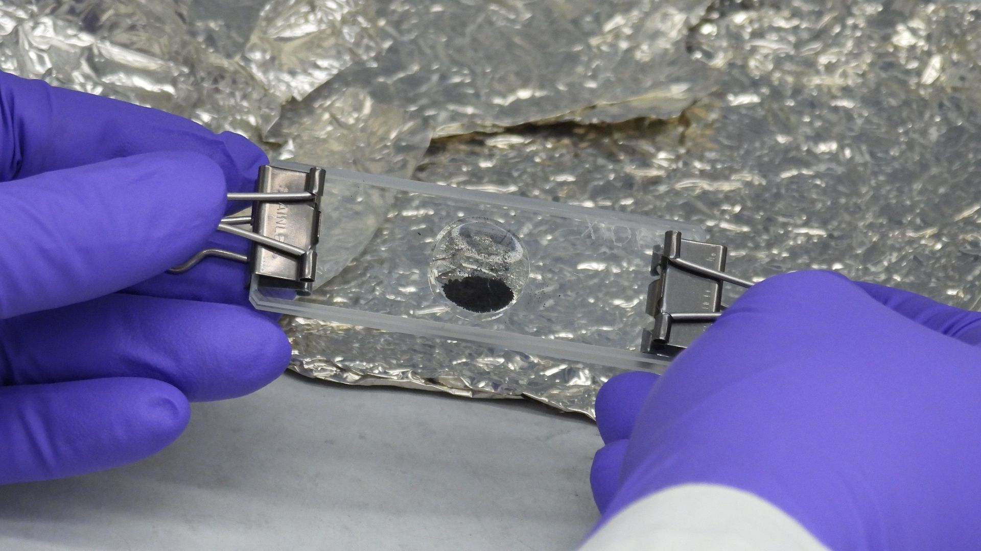 A portion of the asteroid Bennu sample delivered to Earth by NASA's OSIRIS-REx (Origins, Spectral Interpretation, Resource Identification, and Security – Regolith Explorer) mission, set into a microscope slide at the agency's Goddard Space Flight Center i
