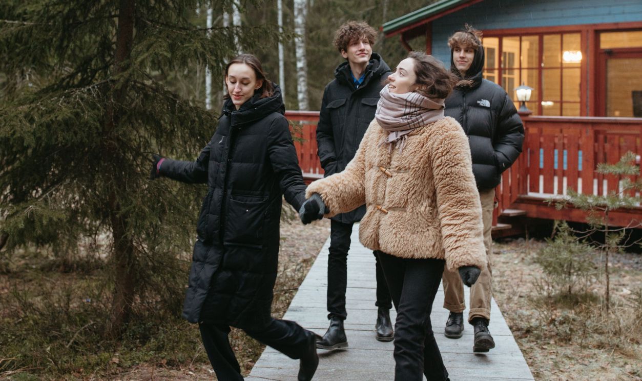 Young adults walk in winter coats along a forest path near a cozy cabin.