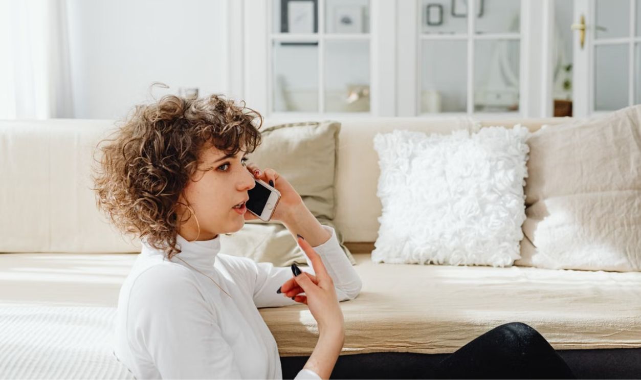 A woman is seated, talking on her phone in a bright, cozy living room.