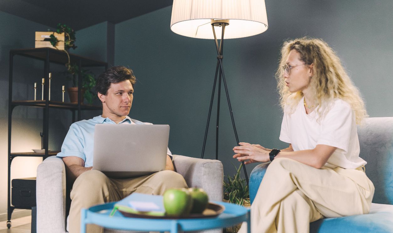 Two adults discuss work while sitting on sofas in a cozy, modern living room setting