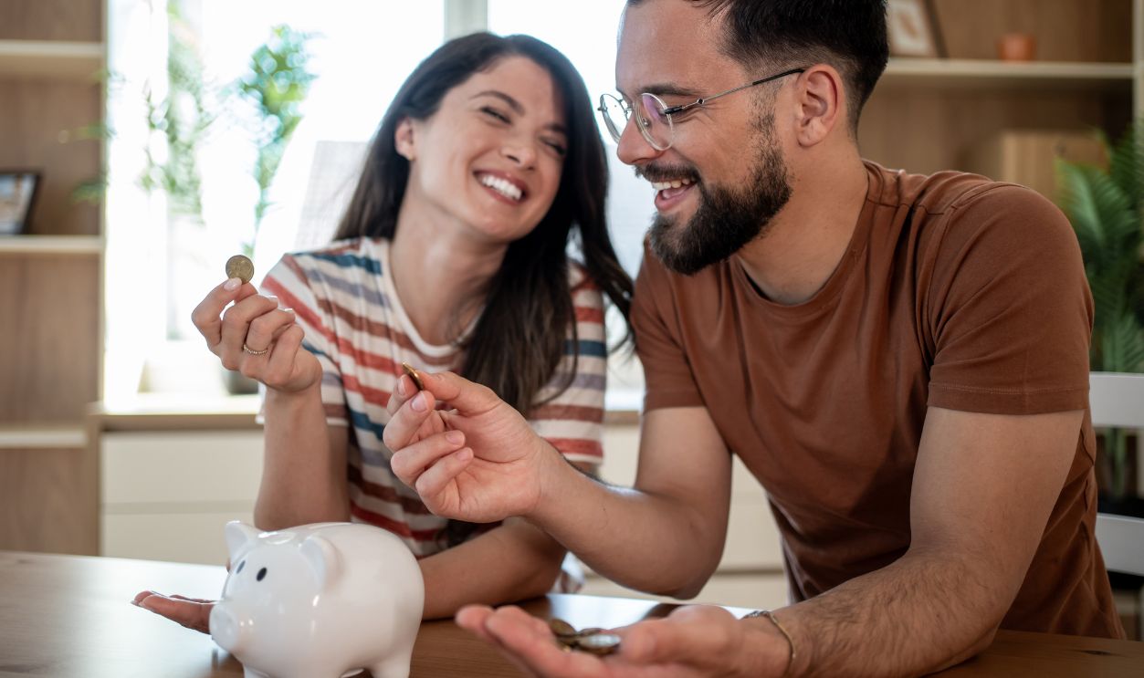Young smiling couple putting coins into piggy bank, saving money for future, managing home budget, making financial plans together, successful investment strategy, happy family life