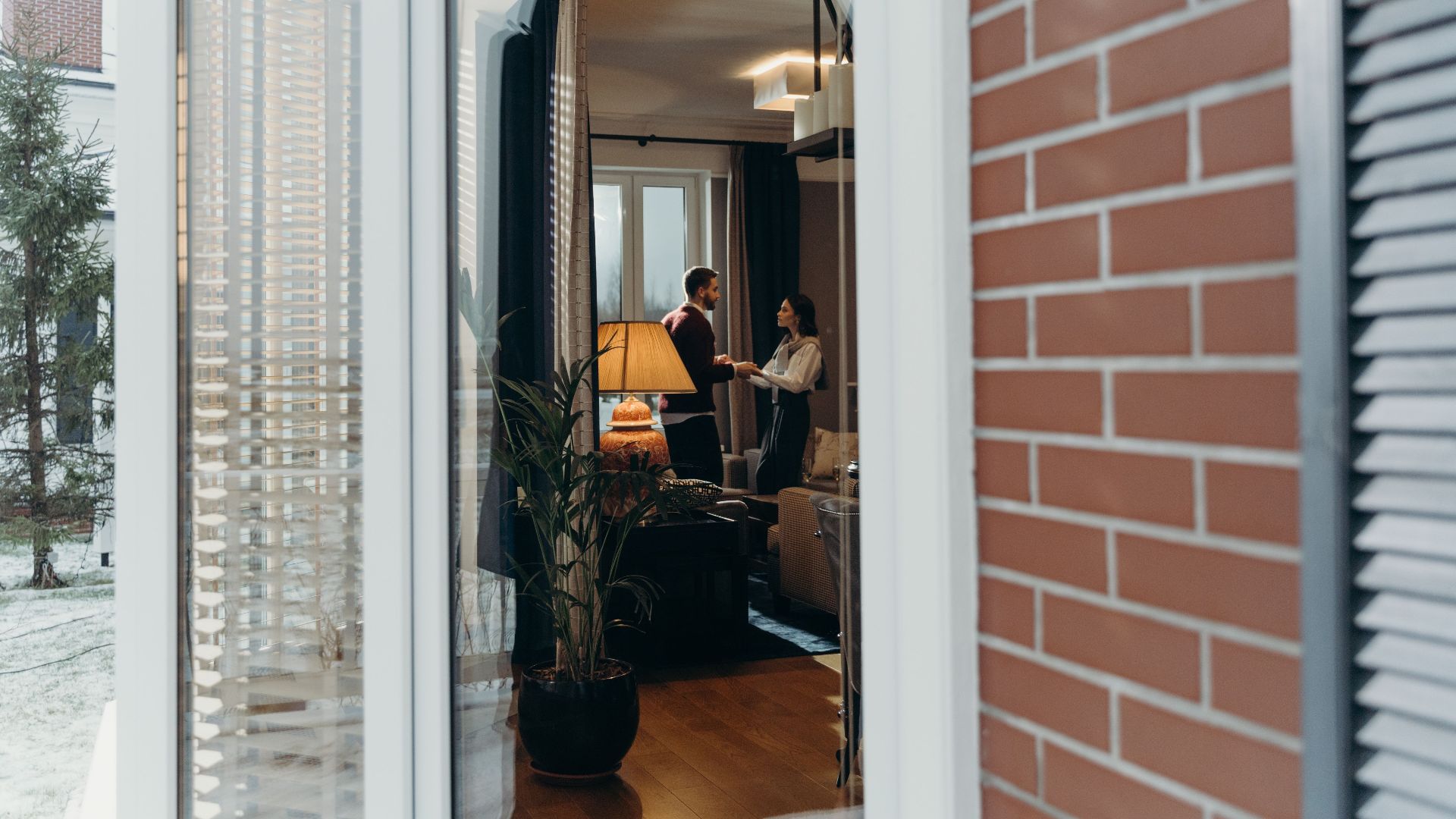 A couple stands in a stylish room seen through a window, holding hands.