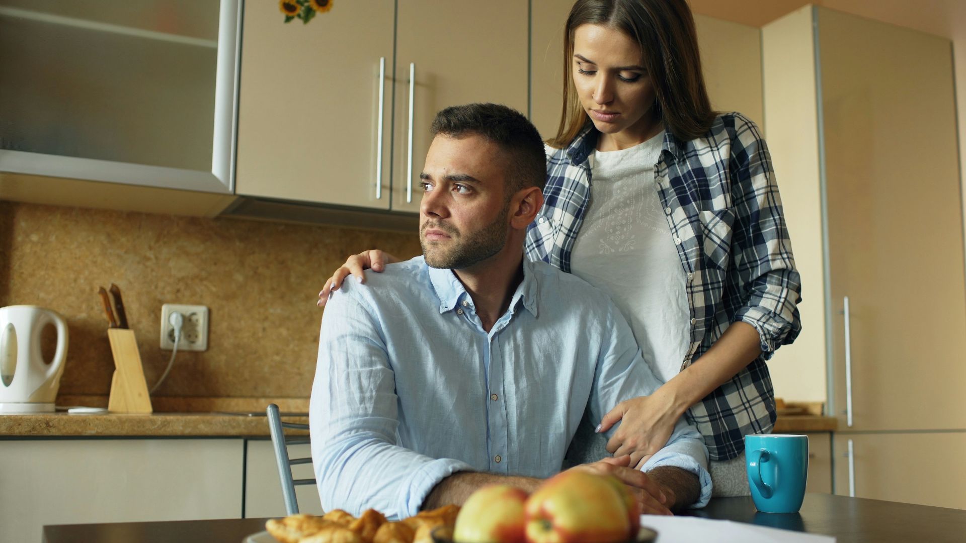 Woman comforts man at kitchen table with food.
