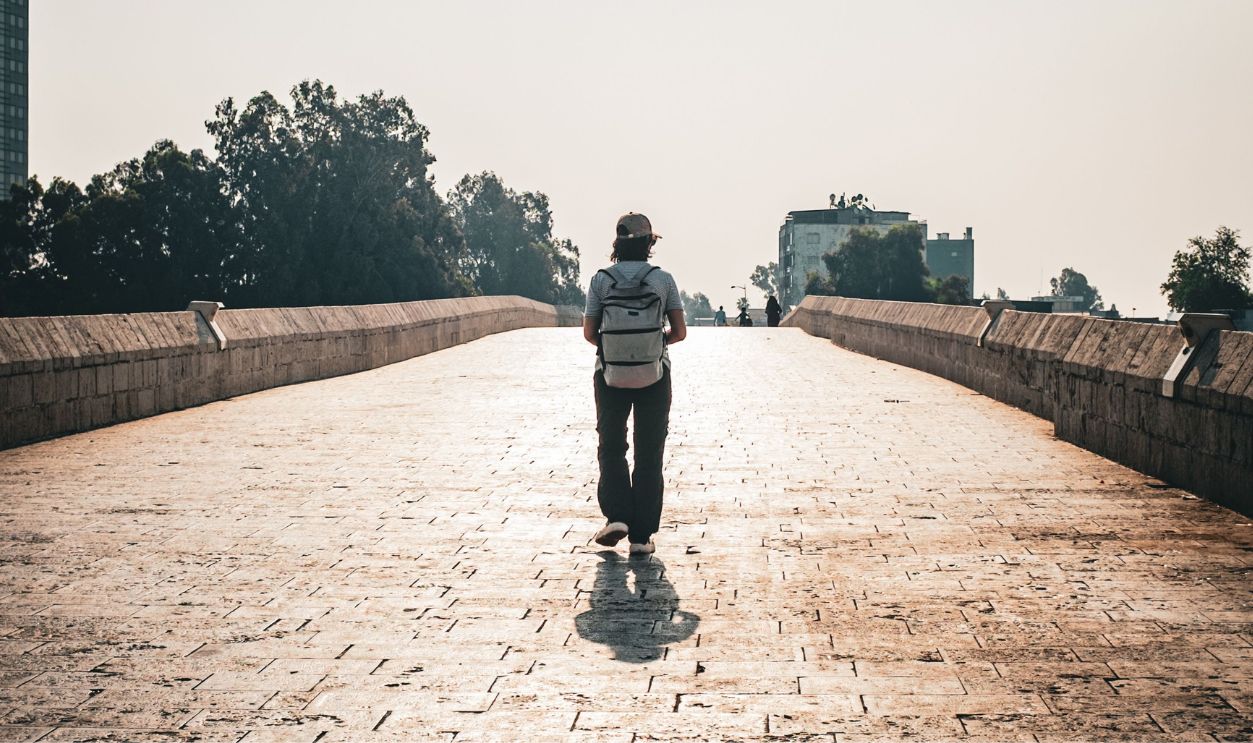 A lone traveler walks on a sunlit stone bridge in an urban setting