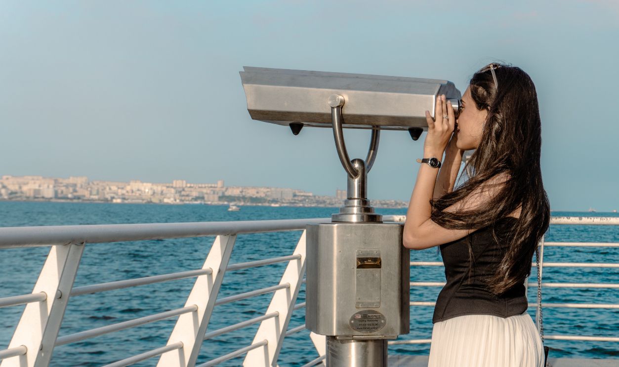A woman uses binoculars on a seaside promenade