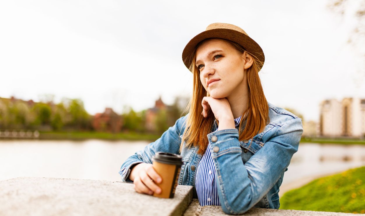 Woman in denim and hat enjoying coffee outdoors with a thoughtful expression