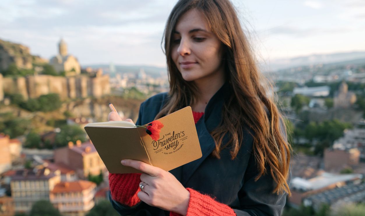 A woman writes in her journal against a backdrop of a historic cityscape.