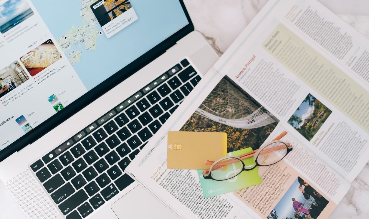 A top view of travel planning essentials on a laptop with a map, credit cards, and a magazine.