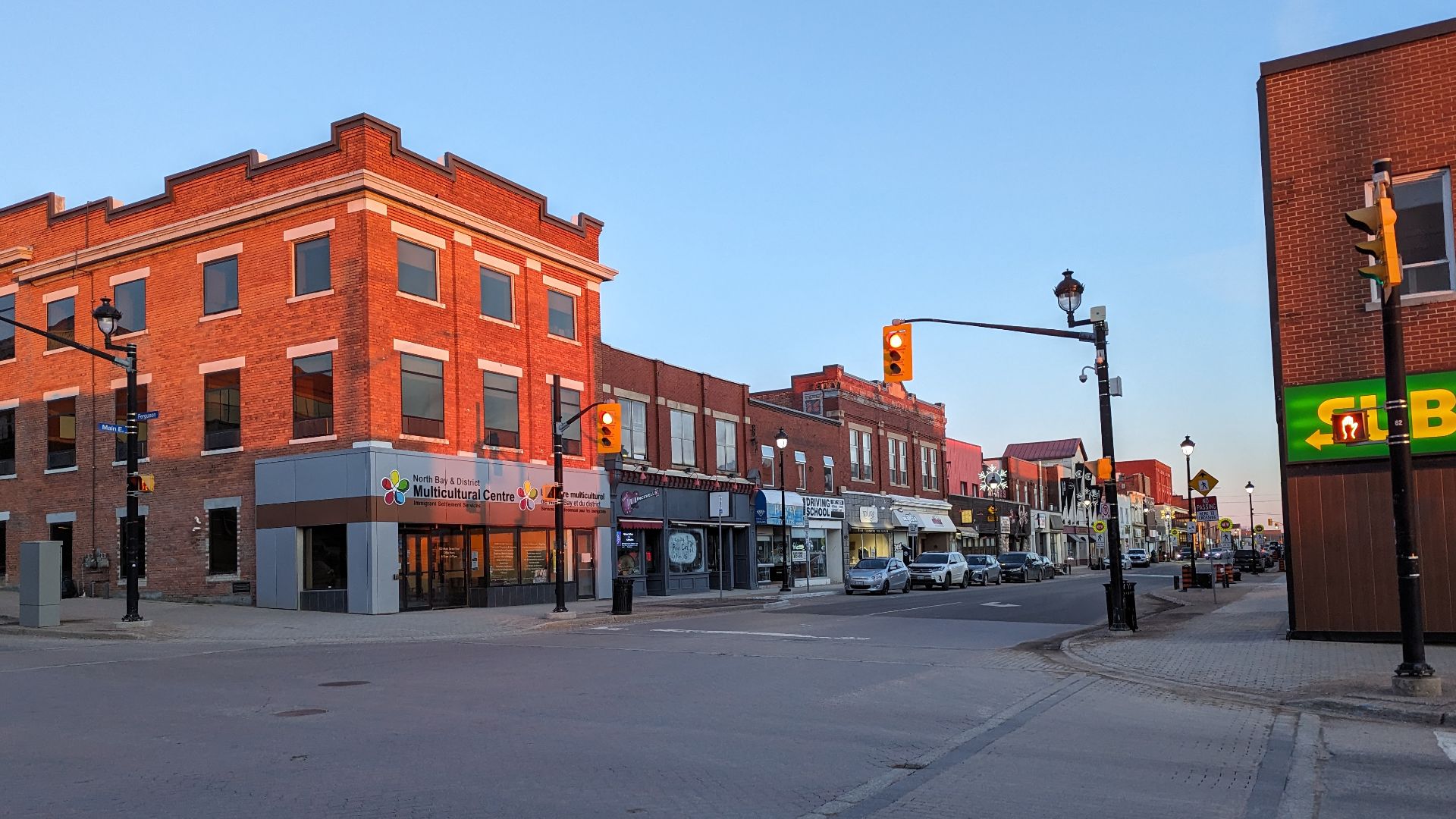 The image shows a quiet, brick-paved intersection with traffic lights and two-story buildings, including a multicultural centre. It's twilight, and there's little traffic or pedestrian activity.