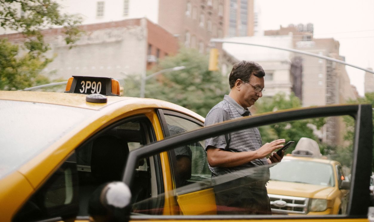 A taxi driver stands by his cab on a bustling city street, using his smartphone