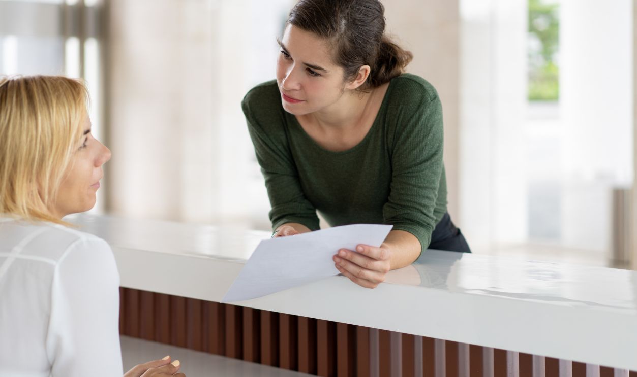Serious female tourist trying to check in. Girl in casual leaning on reception desk, holding paper and talking to receptionist. Hotel and paperwork concept