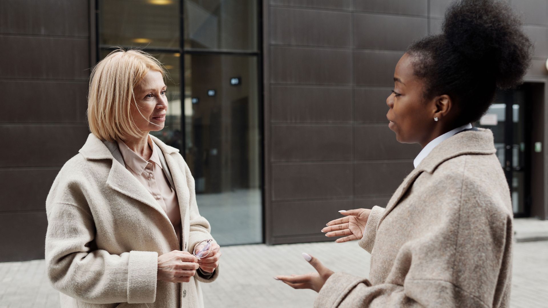 Two businesswomen in coats converse outside a modern office building.