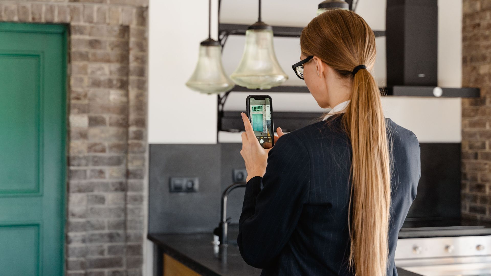 Rear view of a woman with long hair taking a photo with a smartphone in a stylish kitchen.