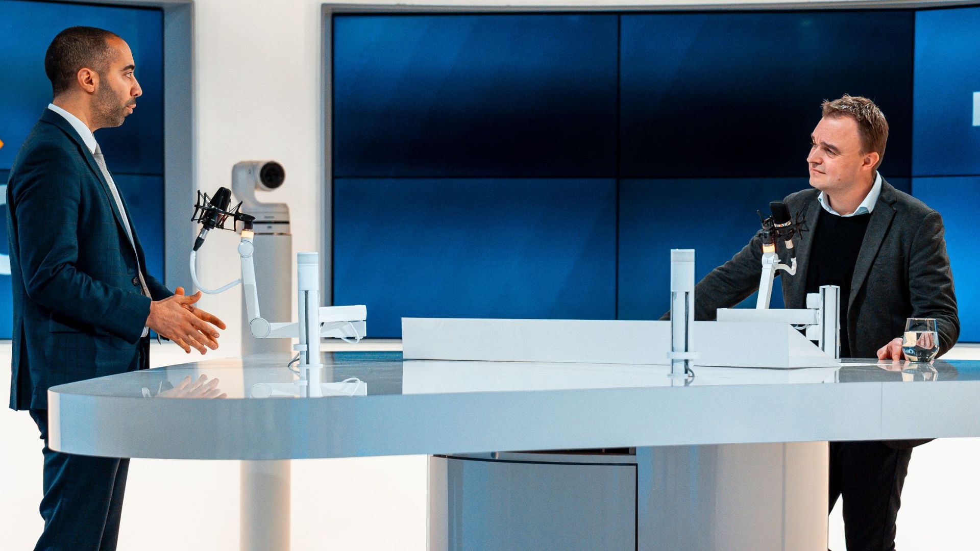Two hosts discussing at a modern TV studio with a dog laying by. Engaging professional setup.