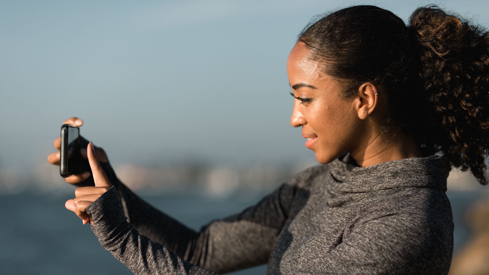 African American woman smiling while taking a photo with her smartphone outdoors near water.