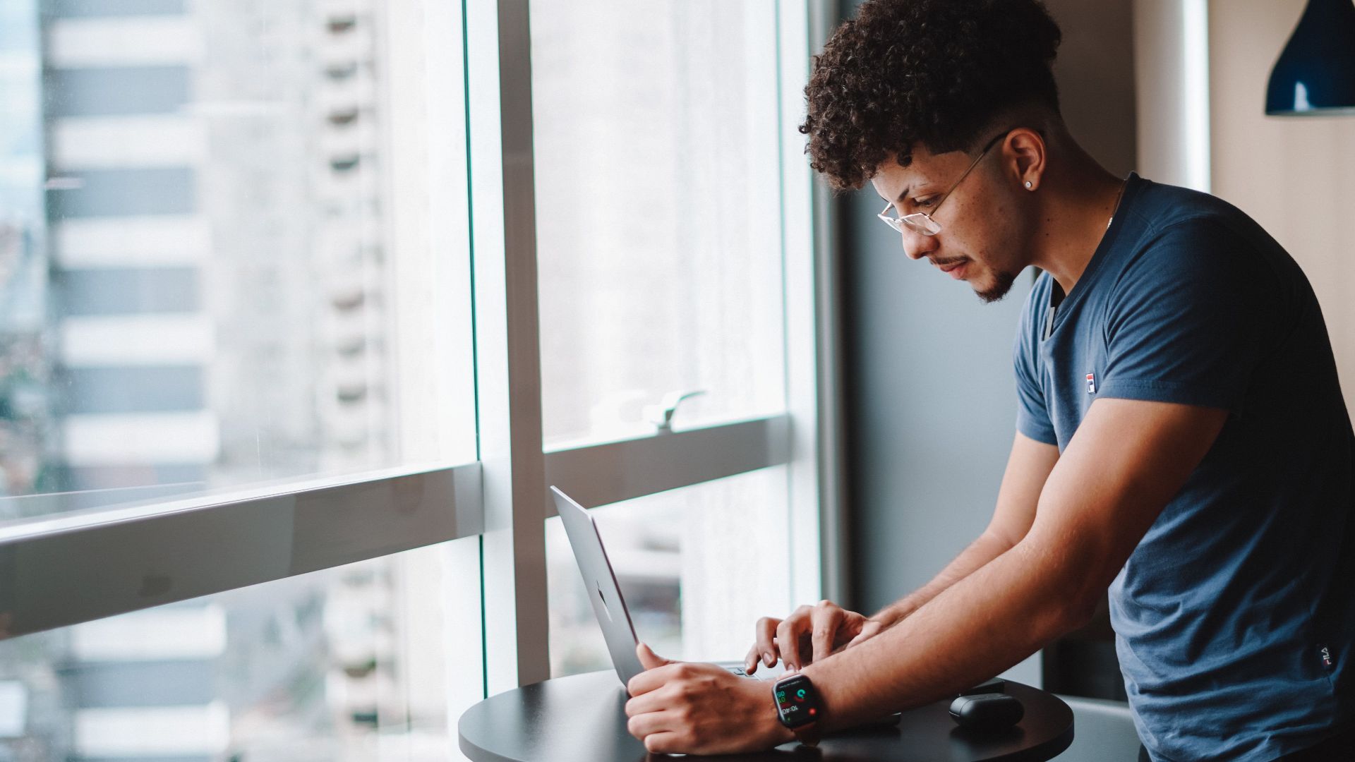 Young man using laptop standing by window in modern office setting.