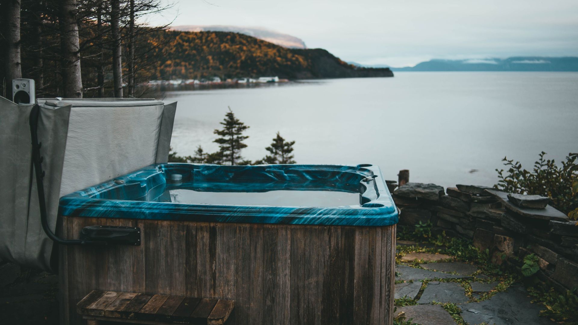 A scenic view of a rustic hot tub by a calm lake surrounded by forest in overcast weather.