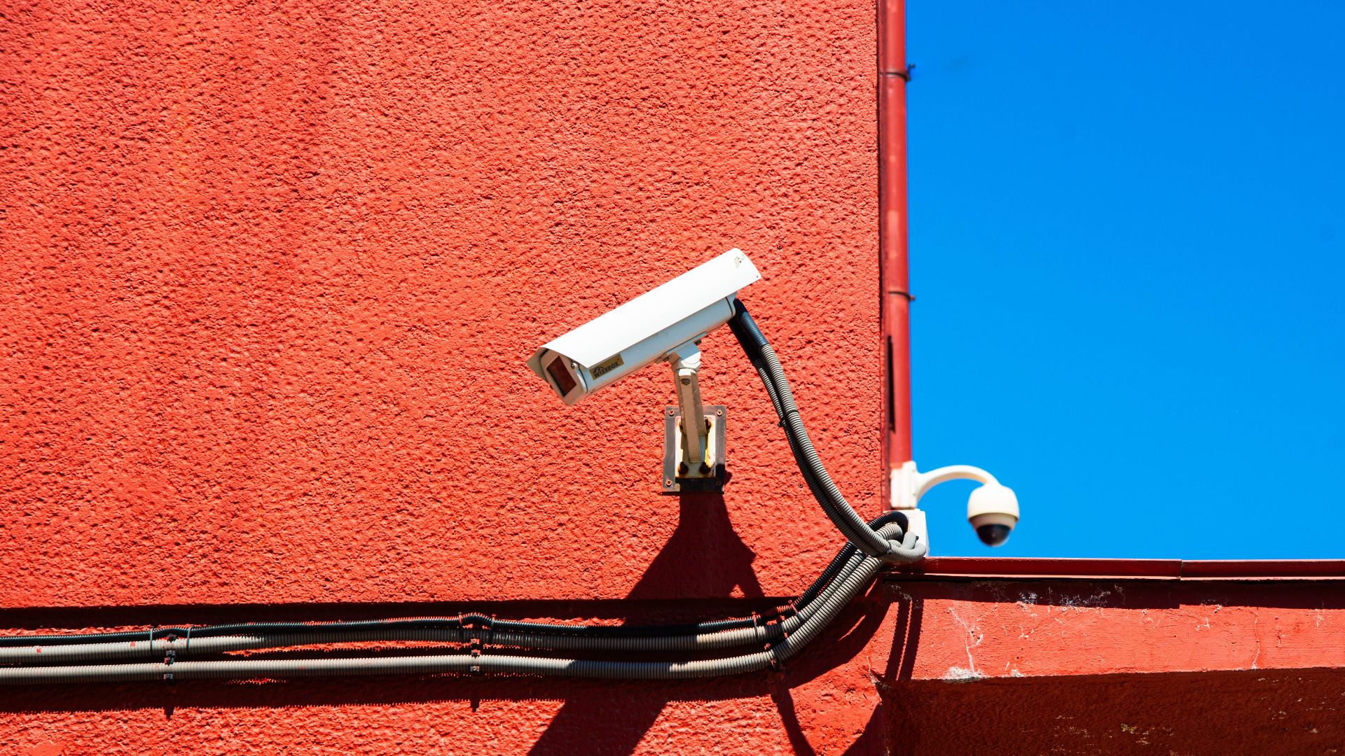 A vivid red wall featuring surveillance cameras and a number sign under a clear blue sky.