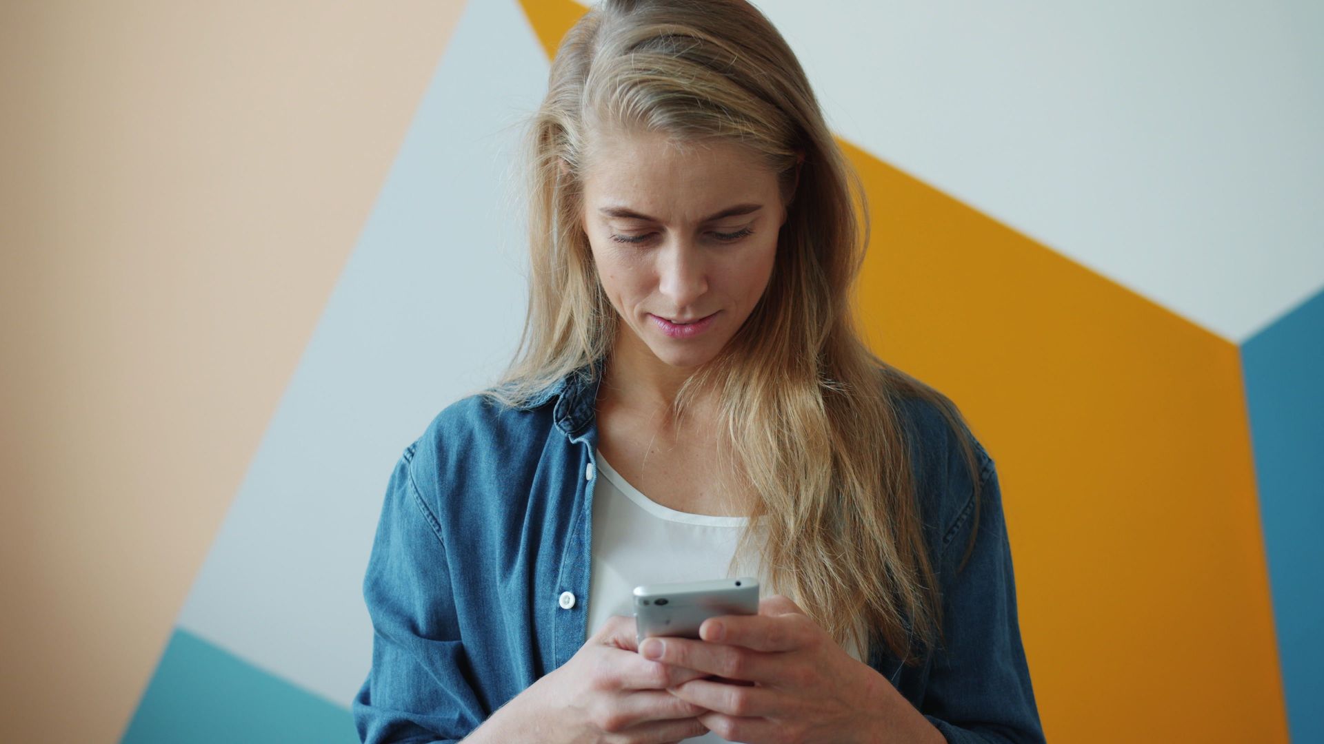 A young woman in a denim shirt uses a smartphone against a geometric colorful wall.