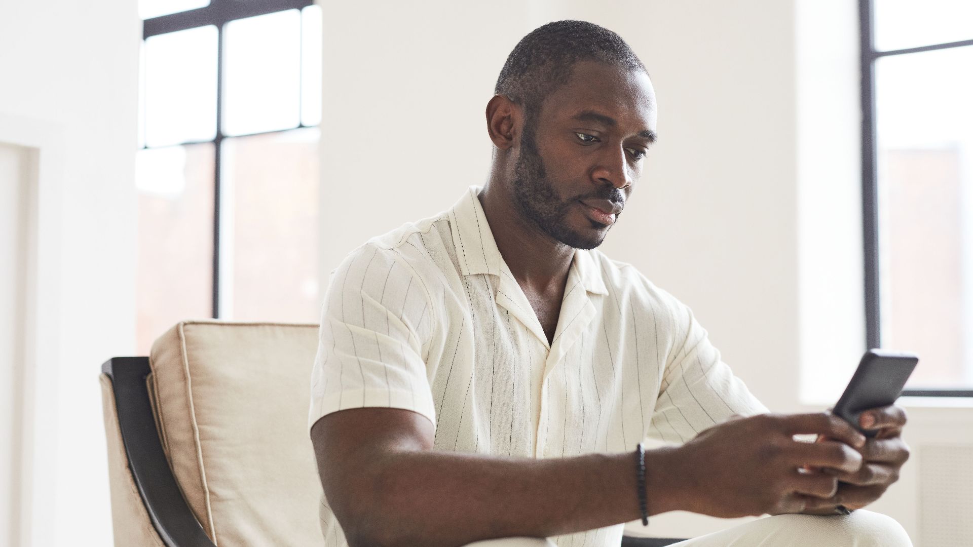 Man with facial hair sitting indoors using smartphone, focusing on screen.