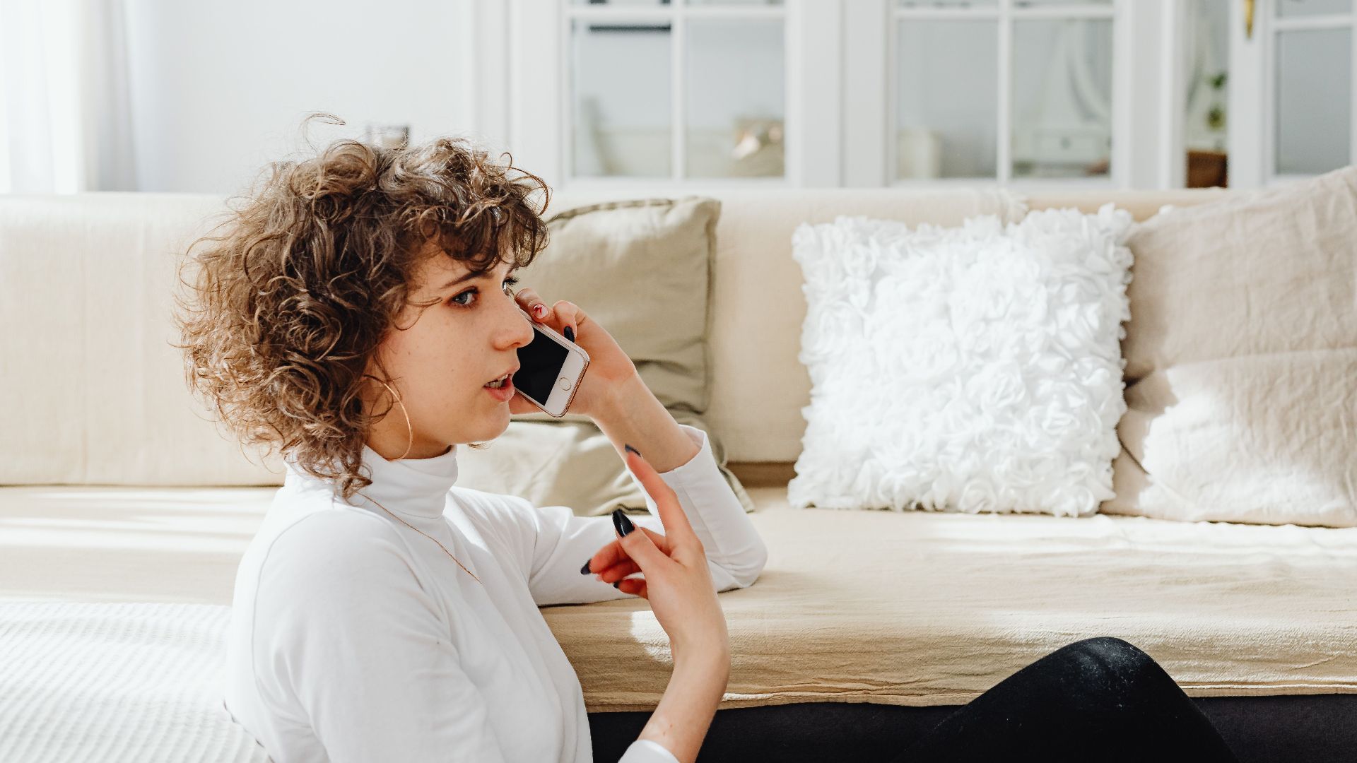 A woman is seated, talking on her phone in a bright, cozy living room.