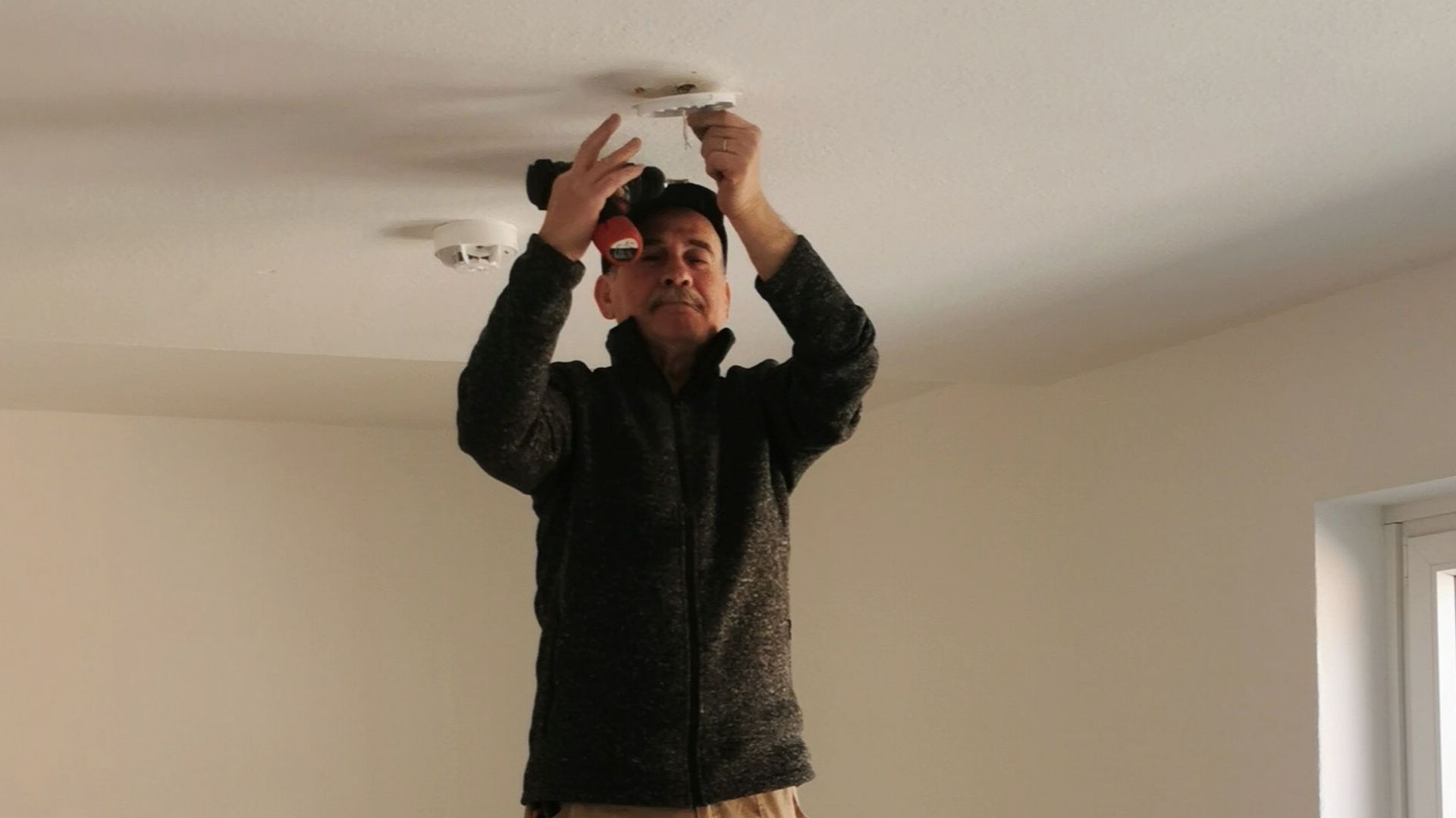 Adult man on ladder fixing a ceiling light in an empty room with beige walls.