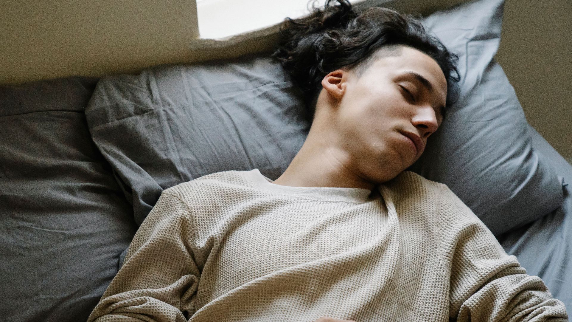 From above of relaxed millennial ethnic male in comfy clothes napping peacefully on soft bed with gray bedclothes