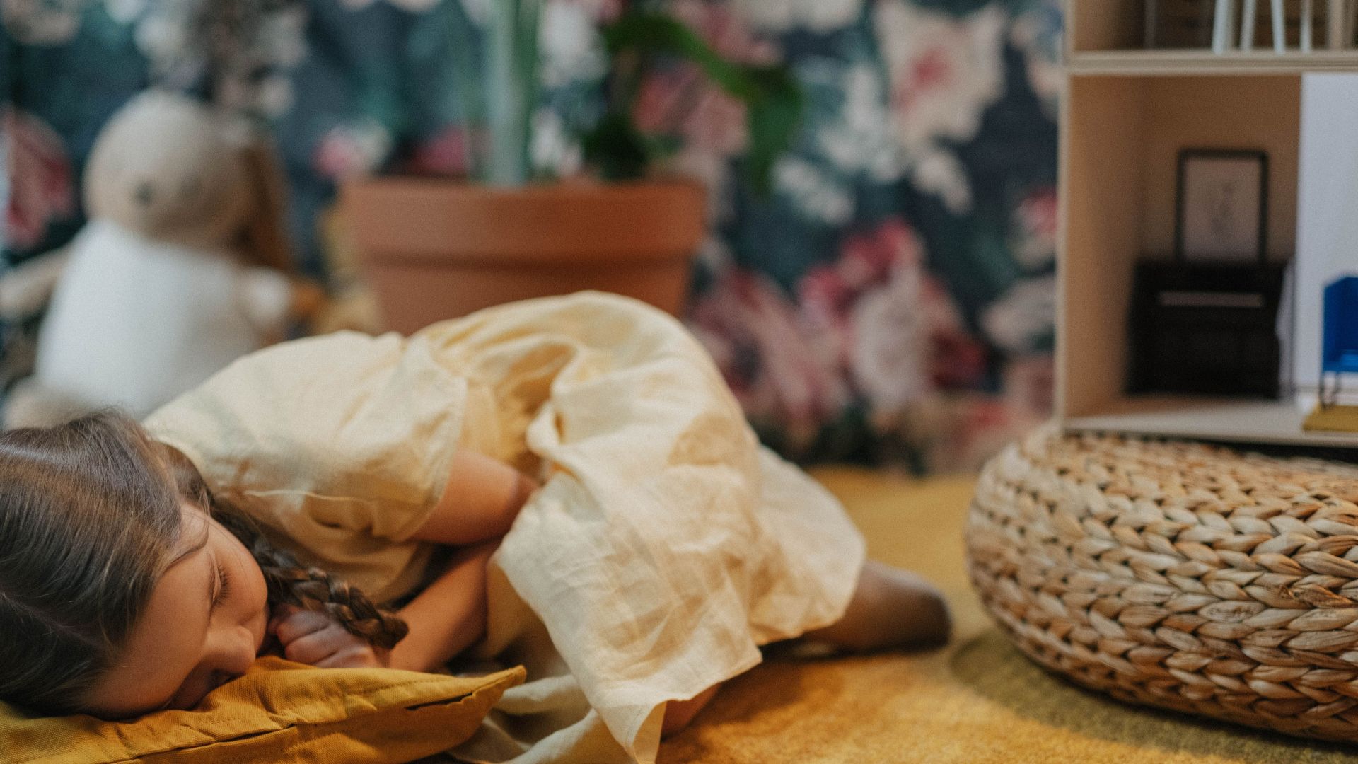 A young girl in a yellow dress peacefully asleep indoors next to a dollhouse and wicker ottoman.