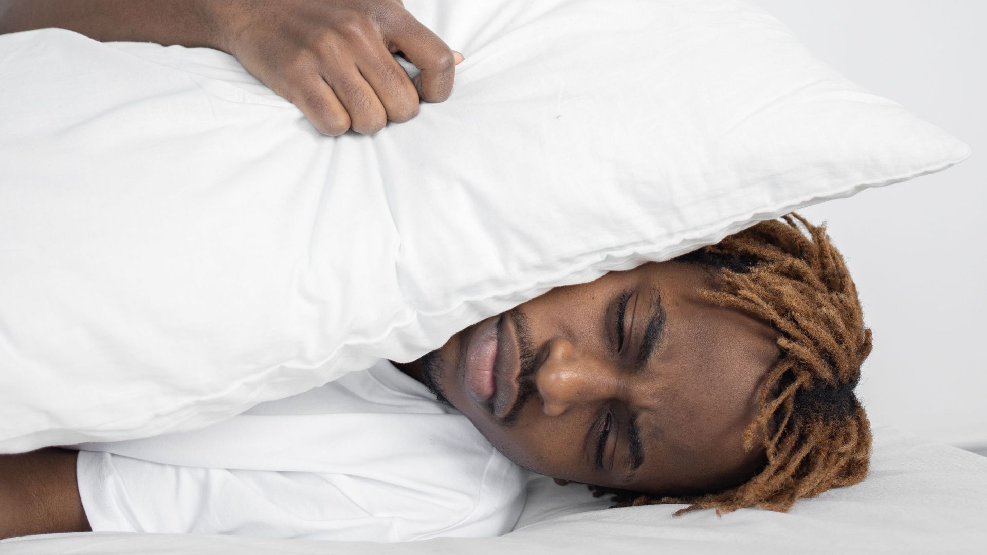 A young man lies in a bed indoors with a pillow covering his face, expressing emotion.