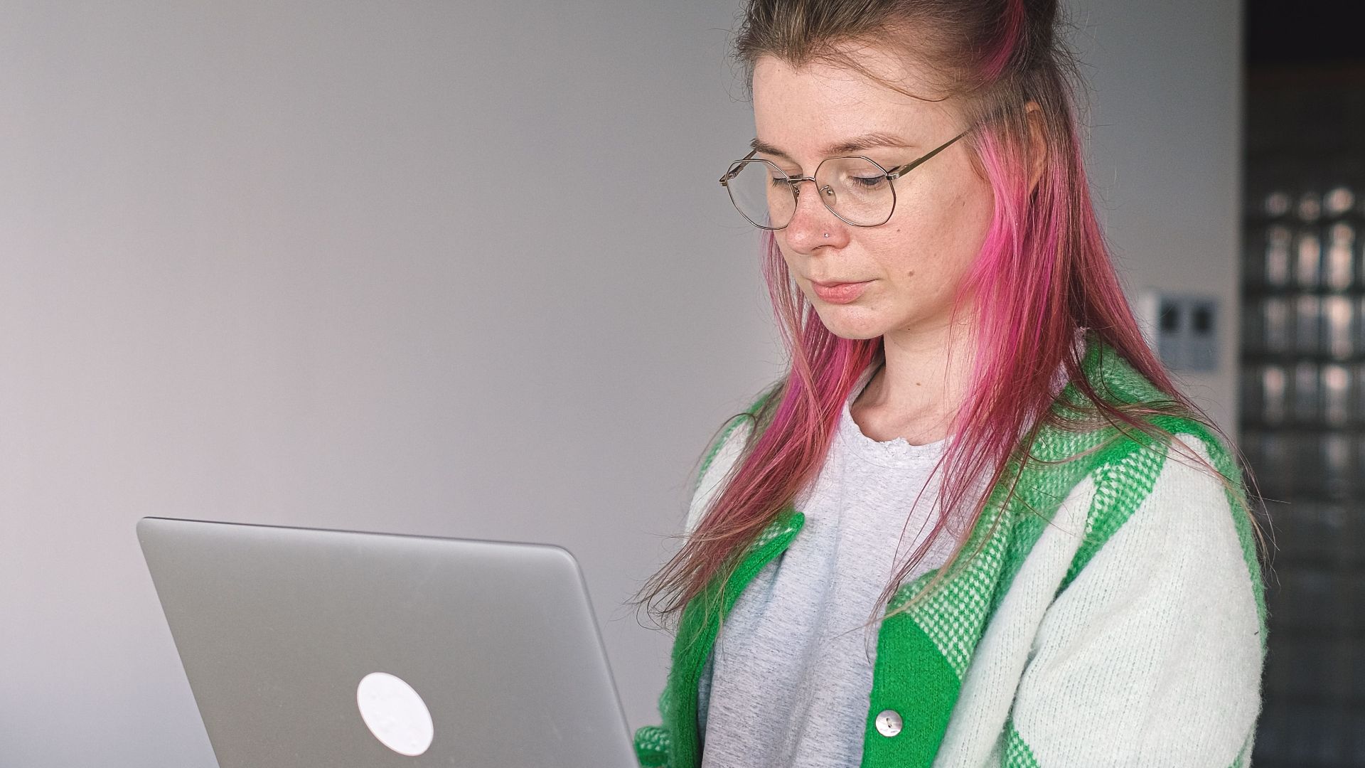 A woman with pink hair is focused on her laptop in a stylish indoor setting, showcasing modern work culture.