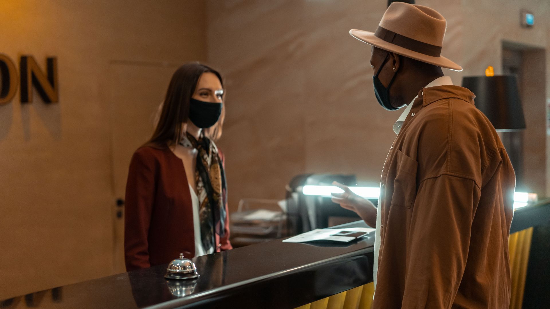 A man at a hotel reception talking to a staff member. Both wear face masks.